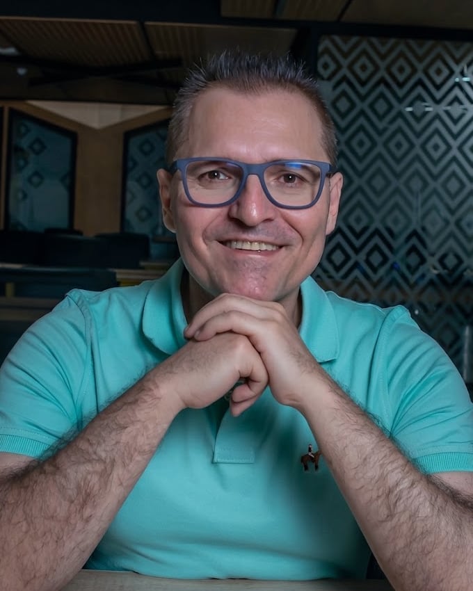 Carlos Rendón smiling at a desk in an open-plan office, hands clasped under his chin.